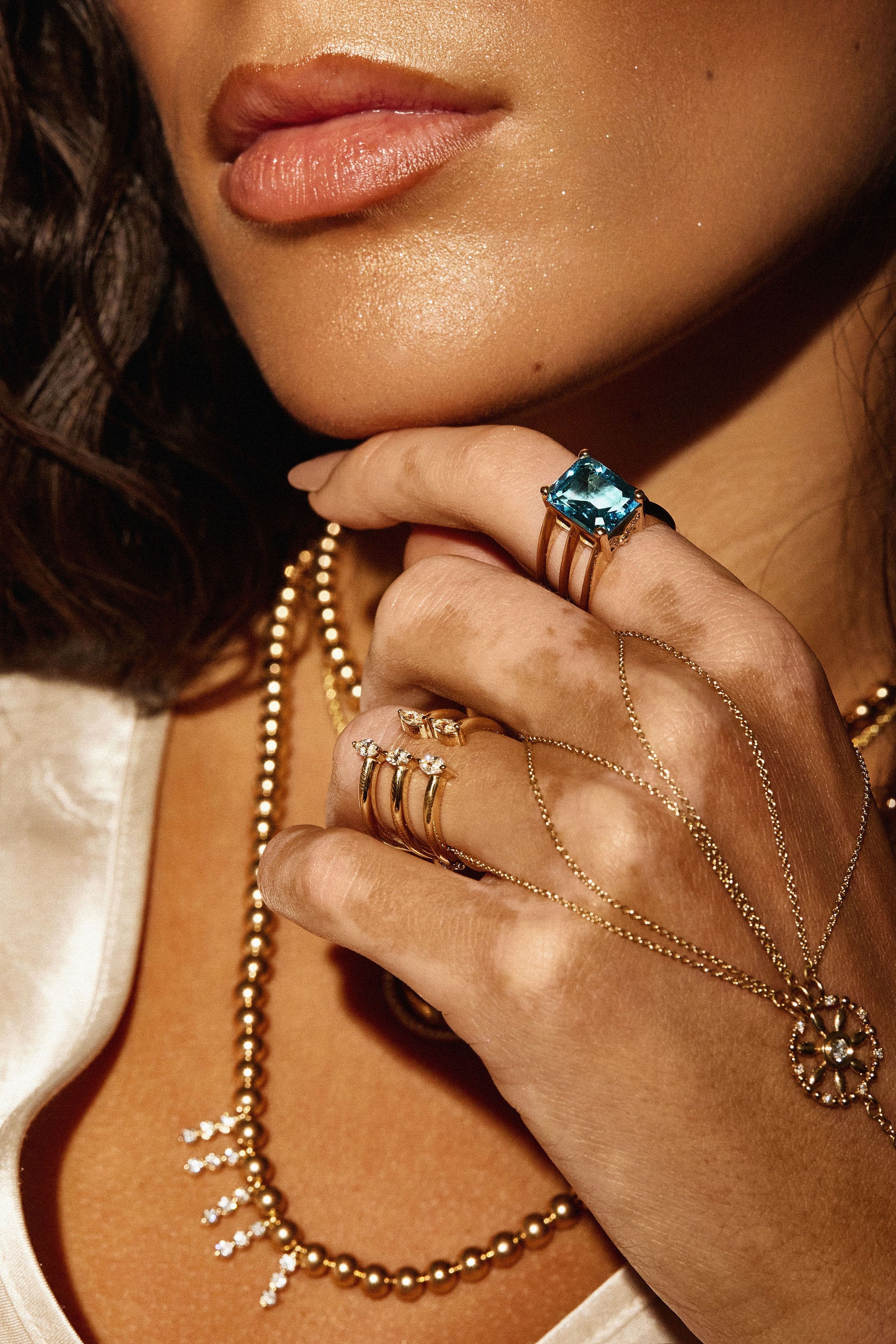 Close-up of a woman wearing multiple 14K yellow gold rings and necklaces. One of the rings is the Nijma M Fine Jewelry Blue Ice - Blue Topaz Triple Band Yellow Gold Ring, featuring a large blue topaz gemstone, while another has a delicate butterfly design. She also wears layered gold necklaces with one featuring a star-shaped pendant adorned with natural diamonds.