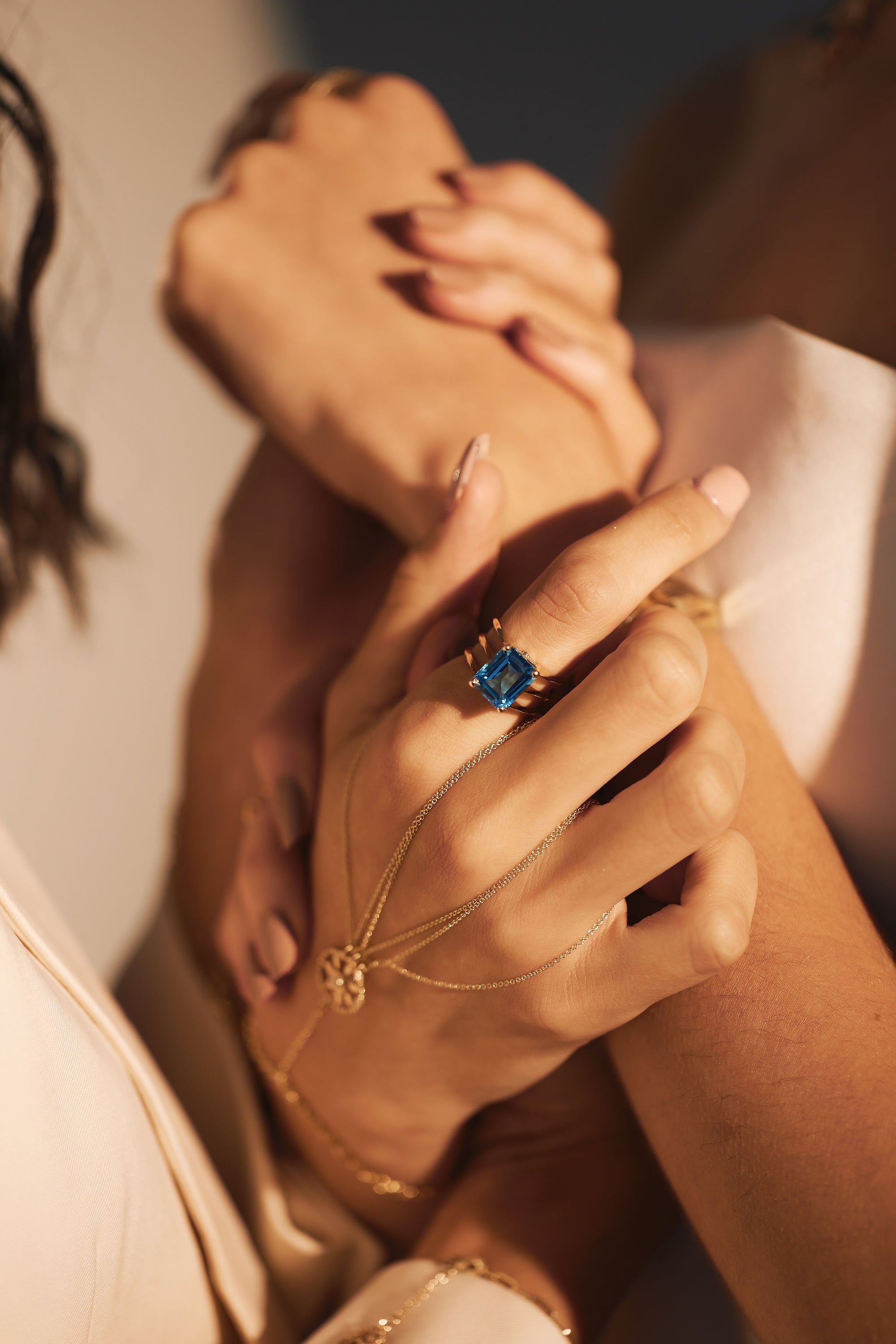 Close-up of intertwined hands, showcasing elegant jewelry. One hand features the stunning Blue Ice - Blue Topaz Triple Band Yellow Gold Ring by Nijma M Fine Jewelry and a delicate 14K yellow gold chain bracelet. The image captures a sense of intimacy and connection, highlighting the beautifully manicured nails and sophisticated accessories adorned with natural diamonds.