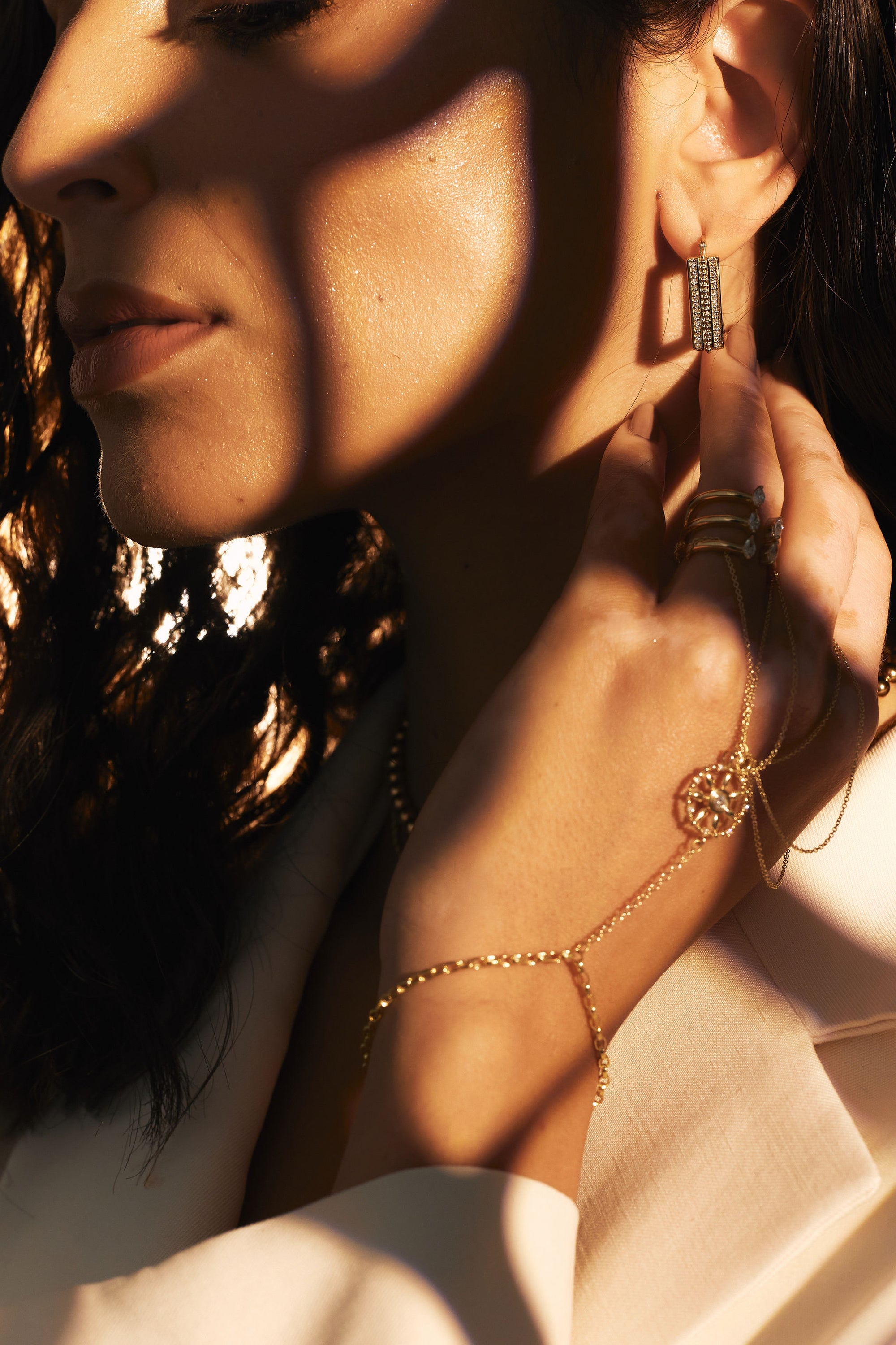 Close-up of a woman showcasing her jewelry. She wears an ear stack with Nijma M Fine Jewelry&#39;s Diamond Plaza - Square Hoop Diamond Earrings, a delicate necklace with a circular pendant, several rings, and a bracelet-ring chain. The lighting casts dramatic shadows on her face and hand, highlighting the shimmer of her natural diamond accessories.