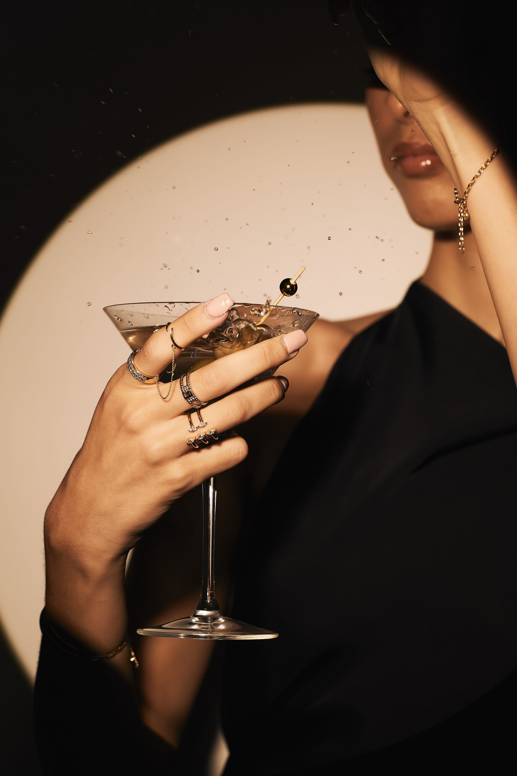 A woman adorned with Nijma M Fine Jewelry's The Double - Chained Yellow Gold Diamond Band and Knuckle Rings, along with dainty gold chains and a bracelet, holds a martini glass garnished with an olive. She is dressed in a sleek black outfit, strikingly highlighted by a spotlight that casts a shadow, drawing attention to the glass and her elegantly decorated hand.