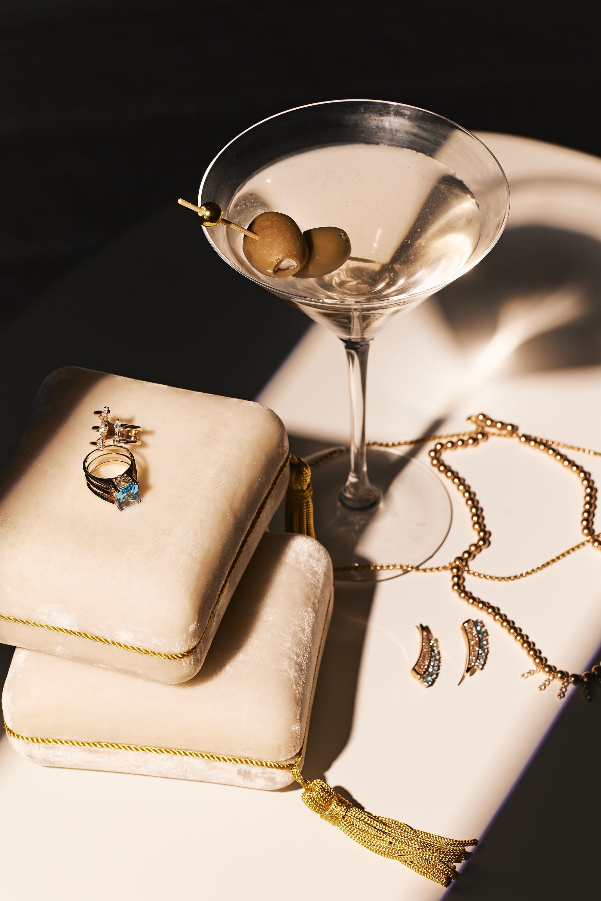 A martini glass with olives sits on a surface next to the Jewelry Pochette Case by Nijma M Fine Jewelry. The case, topped with rings and featuring gold tassel detailing, is surrounded by gold necklaces and earrings adorned with diamonds. Sunlight casts delicate shadows, enhancing the luxurious ambiance.