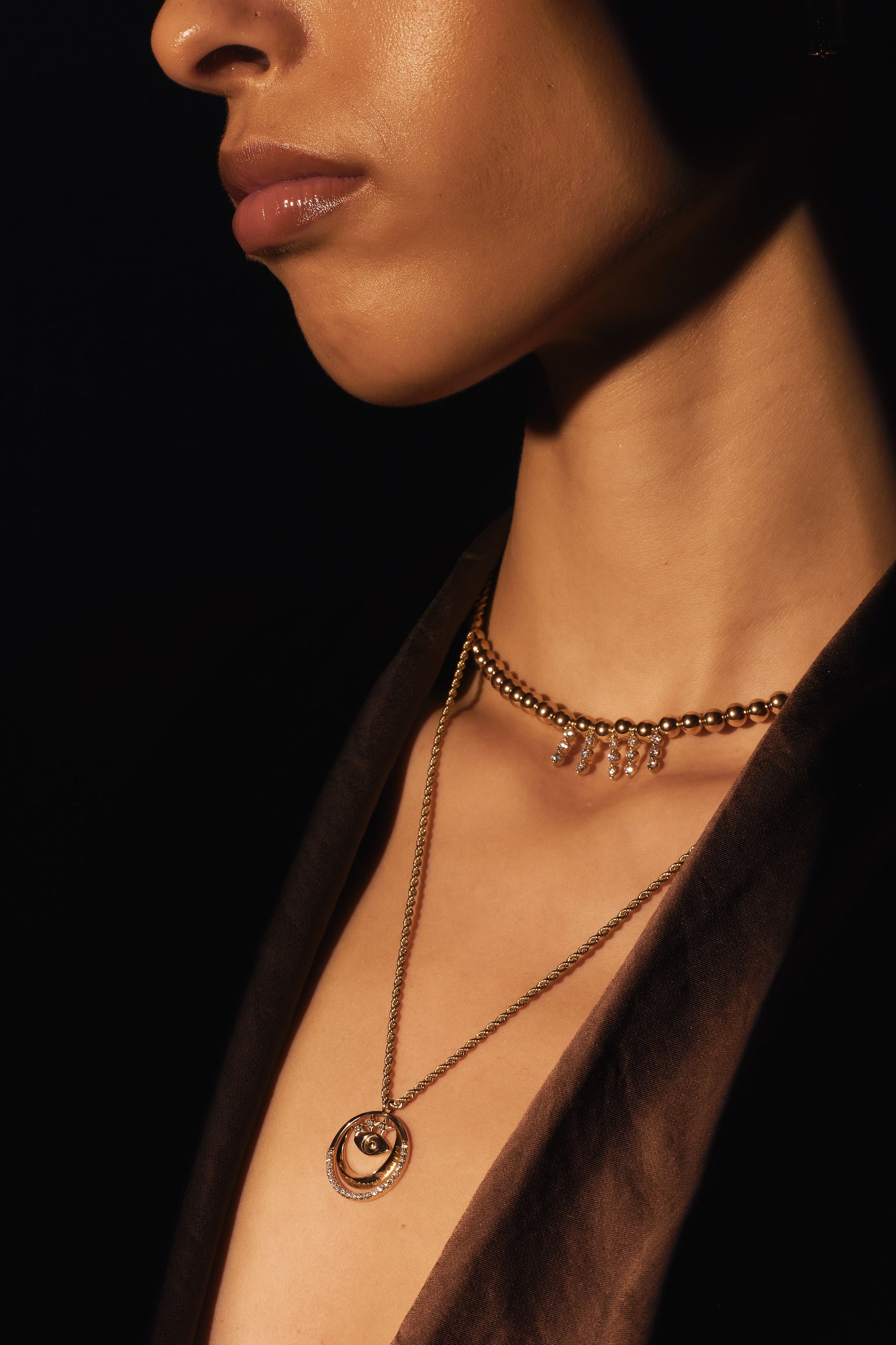 Close-up of a person's neck and lower face, highlighting two necklaces: one is the "Sticks and Gold Stones - Yellow Gold Beads and Diamond Necklace" from Nijma M Fine Jewelry, featuring vibrant gold beads with small dangling charms, while the other is a longer chain with a pendant showcasing two interlocking rings. The low lighting accentuates both the jewelry and the skin's texture.