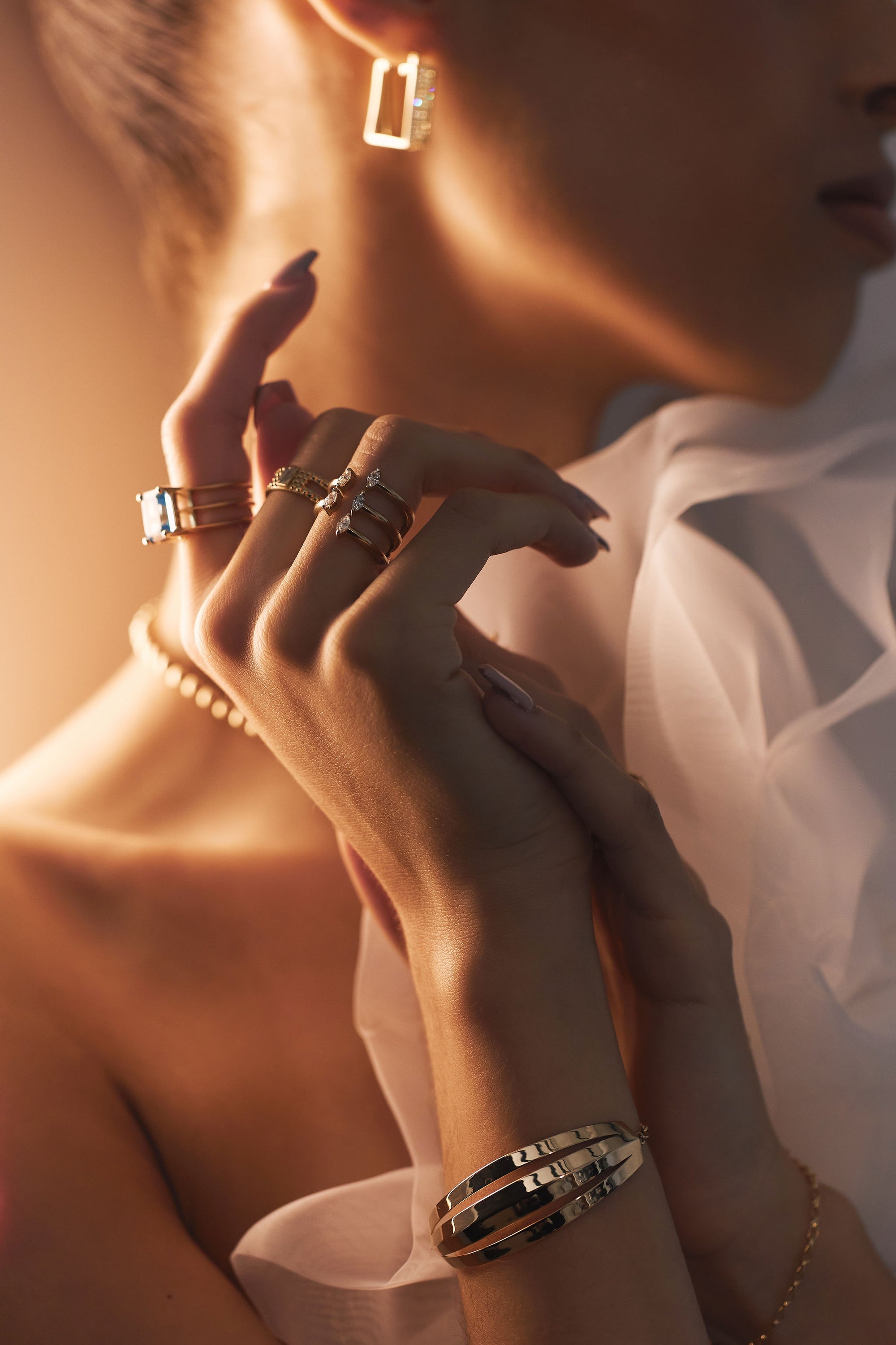 A woman adorned with elegant jewelry, including geometric earrings, multiple rings, a pearl necklace featuring an eye-catching sun charm, and the "Gold Bar - Bar and Chain Yellow Gold Bracelet" from Nijma M Fine Jewelry. She is posed with her hands gracefully touching near her face, wearing a white ruffled garment illuminated by soft, warm light.