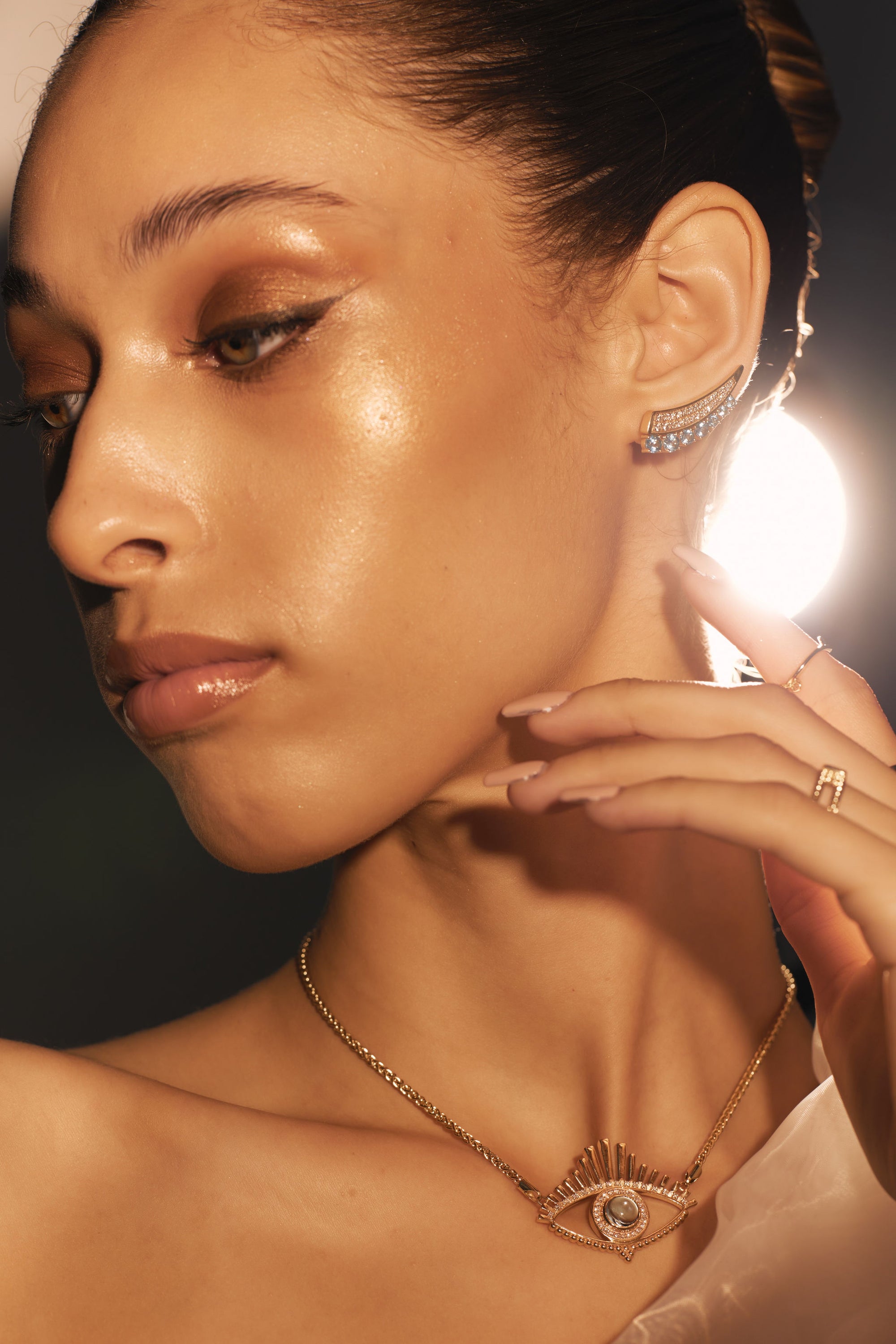 A woman with glowing skin wearing elegant makeup looks to the side, showcasing intricate yellow gold jewelry by Nijma M Fine Jewelry. She has The High Life - Blue Topaz and Diamond Yellow Gold Climber Earrings and a necklace with an eye motif. She lightly touches her ear with her manicured hand, revealing a ring featuring natural diamonds on her finger.