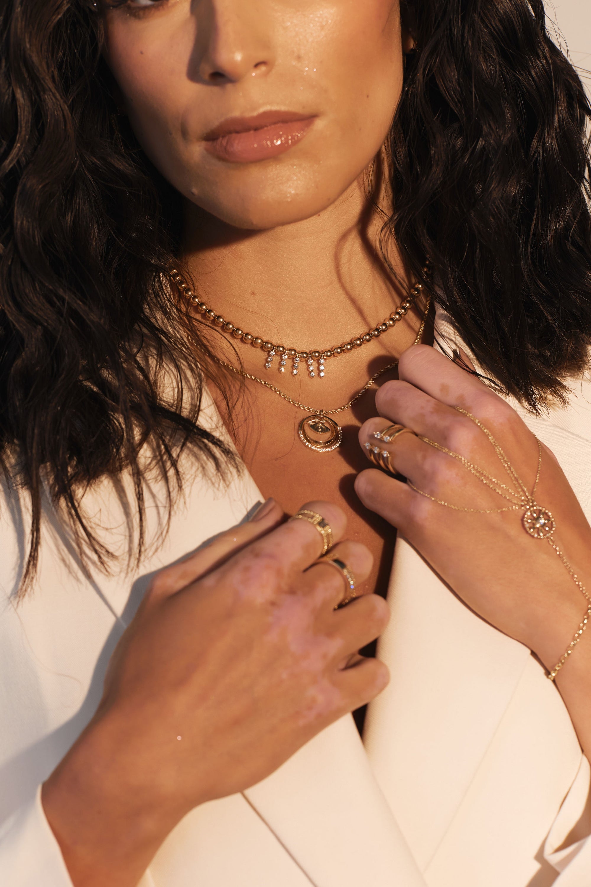 A woman with wavy dark hair wears a white blazer, multiple gold necklaces, and several gold rings. Her hands are crossed near her chest, showcasing her jewelry. The collection includes delicate chains, a sun charm pendant, and the Jazz Fingers - Yellow Gold Hand Chain from Nijma M Fine Jewelry.